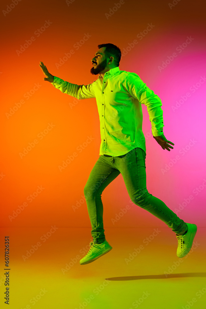 Full-length portrait of handsome bearded man in casual clothes jumping against gradient studio background in neon light. Concept of human emotions, facial expression, lifestyle, fashion
