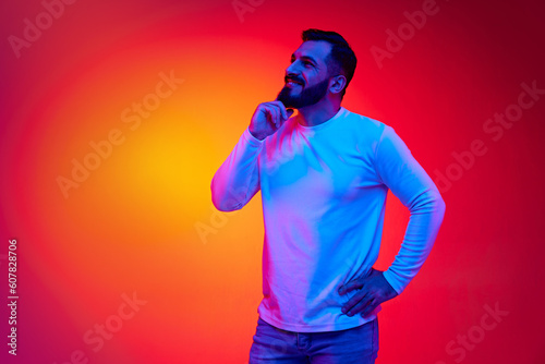 Portrait of handsome bearded man in white shirt posing with smiling, dreaming expression against gradient studio background in neon light. Concept of human emotions, facial expression, lifestyle