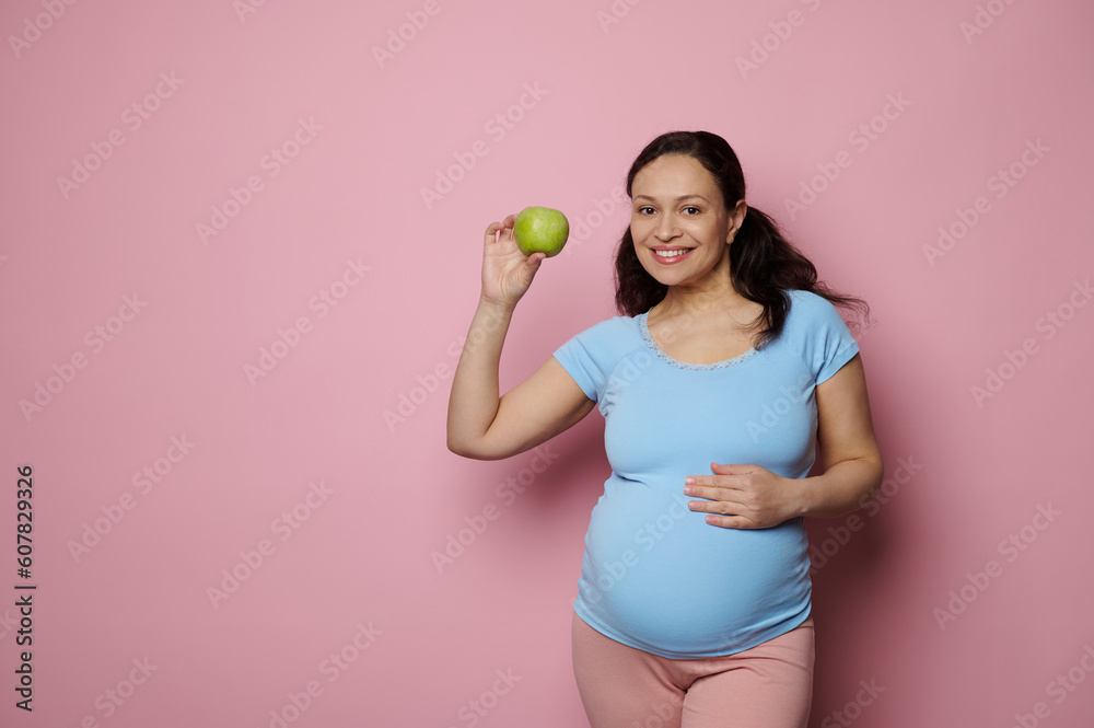 Beautiful pregnant woman wearing a blue casual t-shirt, smiling looking at camera, touching her belly, holding a delicious green apple, isolated on pink background. Healthy nutrition in pregnancy time Beautiful pregnant woman wearing a blue casual t-shirt, smiling looking at camera, touching her belly, holding a delicious green apple, isolated on pink background. Healthy nutrition in pregnancy time