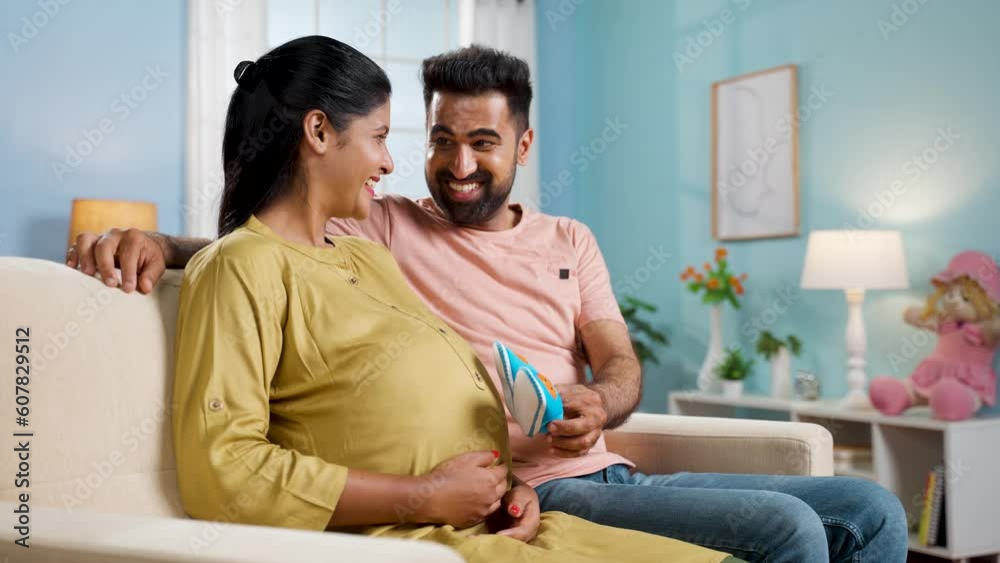 Happy indian playful husband placing baby booties on tummy of pregnant wife at home - concept of maternity preparation, family relationships and parenthood. Happy indian playful husband placing baby booties on tummy of pregnant wife at home - concept of maternity preparation, family relationships and parenthood.