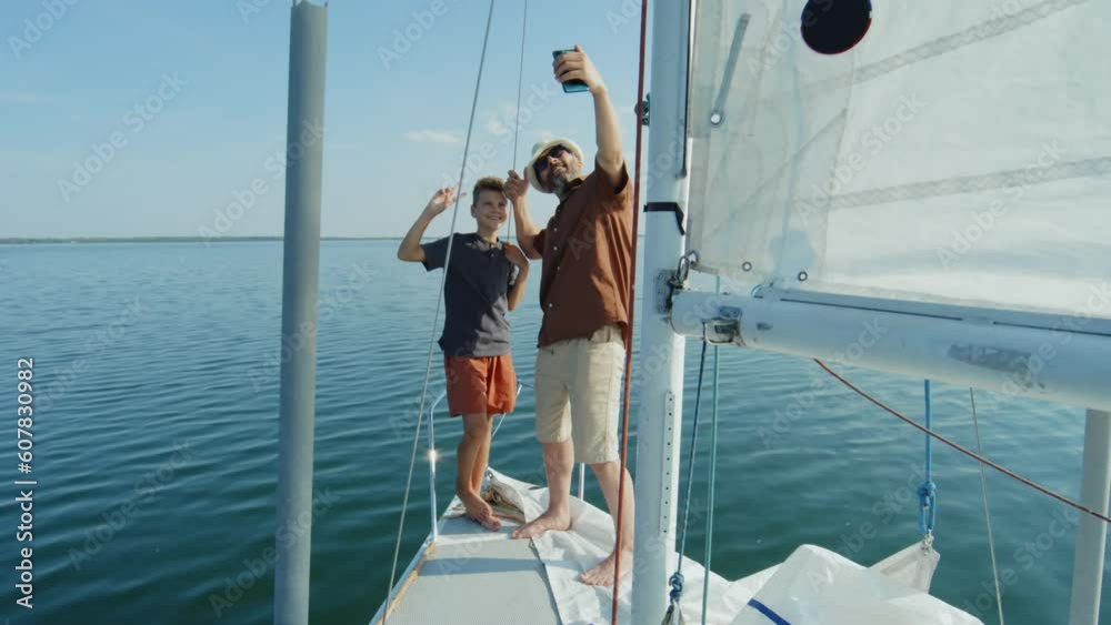 Long shot of modern Caucasian father and his son spending summer day together standing on sailing yacht taking selfie on smartphone Long shot of modern Caucasian father and his son spending summer day together standing on sailing yacht taking selfie on smartphone