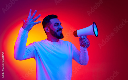 Portrait of excited, happy, smiling man in striped shirt shouting in megaphone against gradient red studio background in neon light. Concept of human emotions, facial expression, lifestyle
