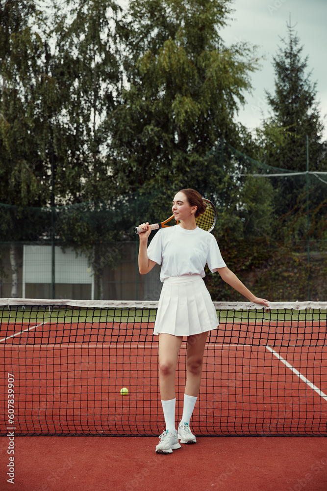 Vertical photo of fashionable girl in white clothing holding tennis racket behind head, posing at tennis net on court. Sports Fashion