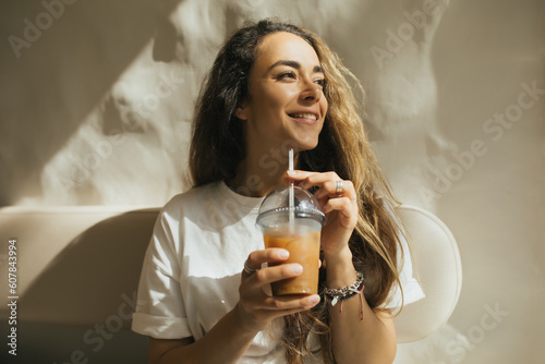 Young woman with long curly hair drinking iced coffee on beige background.