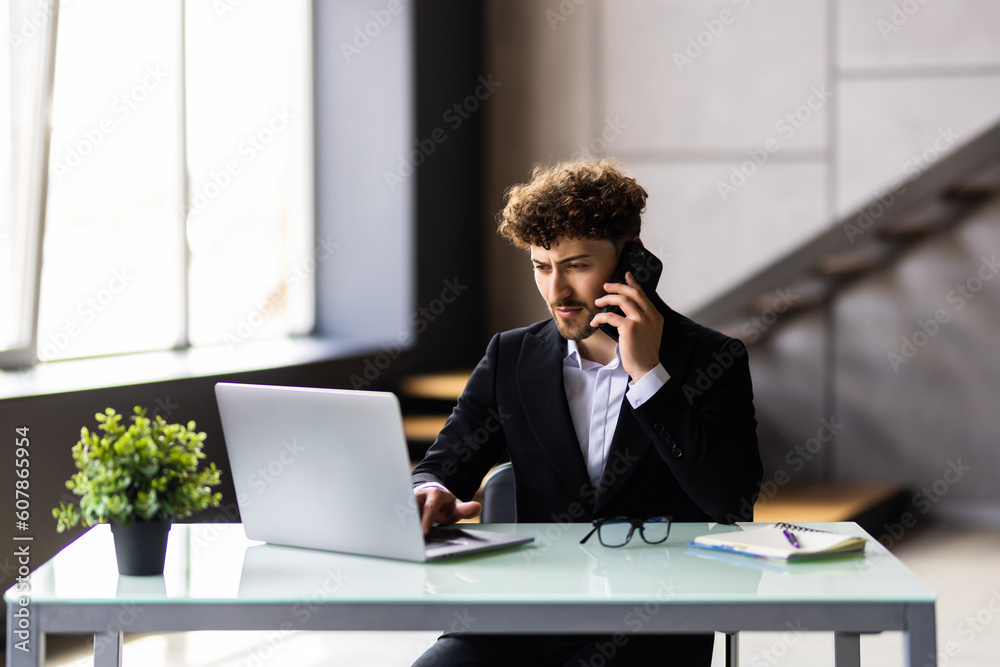 Young man working on laptop and talking on the mobile phone while sitting at his working place in office Young man working on laptop and talking on the mobile phone while sitting at his working place in office