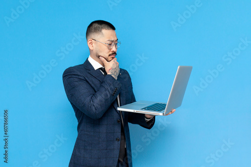 successful pensive asian businessman in suit looks into laptop and thinks on blue isolated background