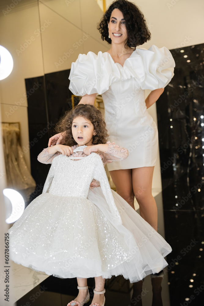 happy middle eastern bride with brunette hair standing in white wedding gown with puff sleeves and ruffles near surprised daughter holding girly dress with tulle skirt near mirror in boutique