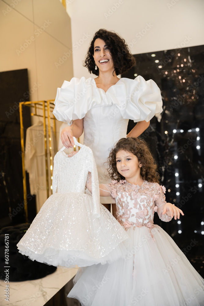 charming middle eastern bride with brunette hair standing in white wedding gown with puff sleeves and ruffles and holding girly dress with tulle skirt near daughter in bridal store charming middle eastern bride with brunette hair standing in white wedding gown with puff sleeves and ruffles and holding girly dress with tulle skirt near daughter in bridal store