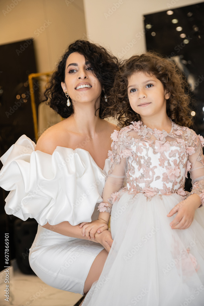 enchanting middle eastern bride with brunette hair in white wedding dress with puff sleeves and ruffles hugging daughter and looking away in bridal store, special occasion enchanting middle eastern bride with brunette hair in white wedding dress with puff sleeves and ruffles hugging daughter and looking away in bridal store, special occasion