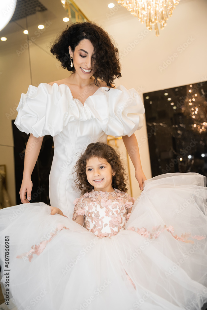 joyful middle eastern little girl in floral attire holding tulle skirt and looking at mirror near bride with wavy hair standing in wedding dress with puff sleeves and ruffles in bridal boutique