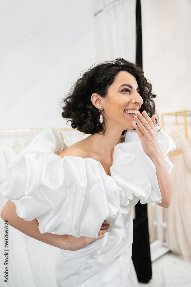 radiant middle eastern bride with brunette wavy hair laughing, covering mouth and posing in trendy wedding dress with puff sleeves and ruffles in bridal salon, fashion radiant middle eastern bride with brunette wavy hair laughing, covering mouth and posing in trendy wedding dress with puff sleeves and ruffles in bridal salon, fashion