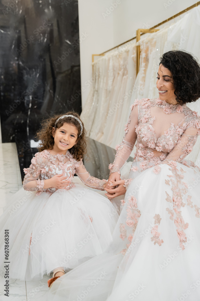 brunette middle eastern woman with wavy hair looking at cheerful girl and smiling near white wedding dresses in bridal salon, floral, mother and daughter, happiness, shopping, wedding day brunette middle eastern woman with wavy hair looking at cheerful girl and smiling near white wedding dresses in bridal salon, floral, mother and daughter, happiness, shopping, wedding day