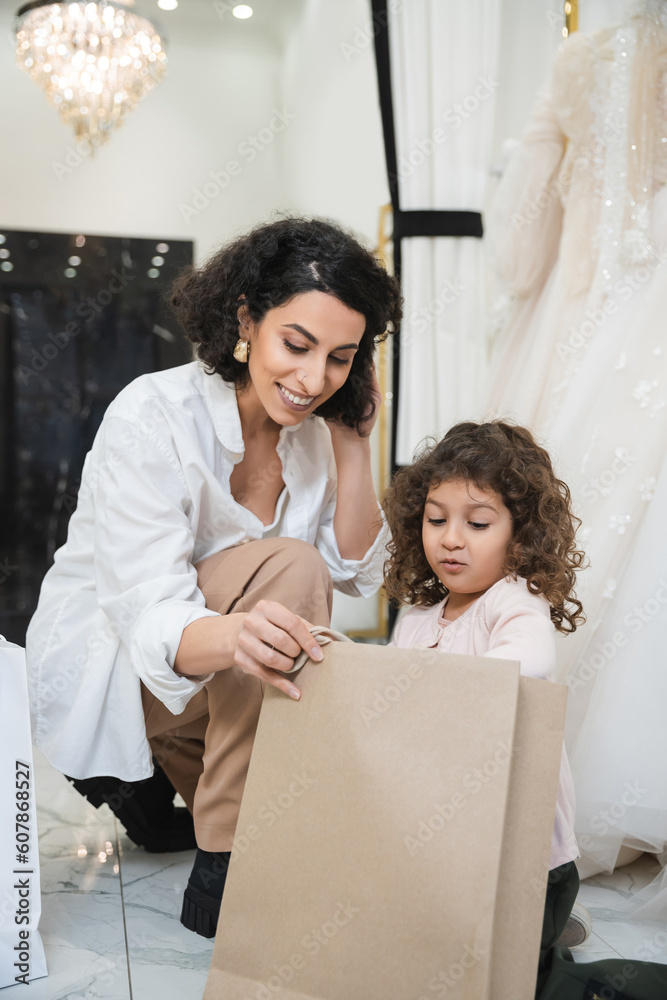cheerful middle eastern woman with brunette hair in white shirt and surprised little girl looking inside of shopping bag near white wedding dresses in bridal salon, mother and daughter cheerful middle eastern woman with brunette hair in white shirt and surprised little girl looking inside of shopping bag near white wedding dresses in bridal salon, mother and daughter