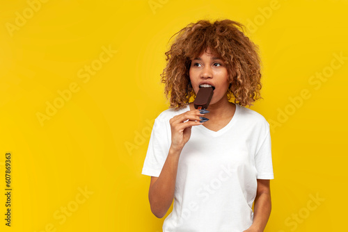 young curly american girl with braces eats chocolate ice cream on yellow isolated background