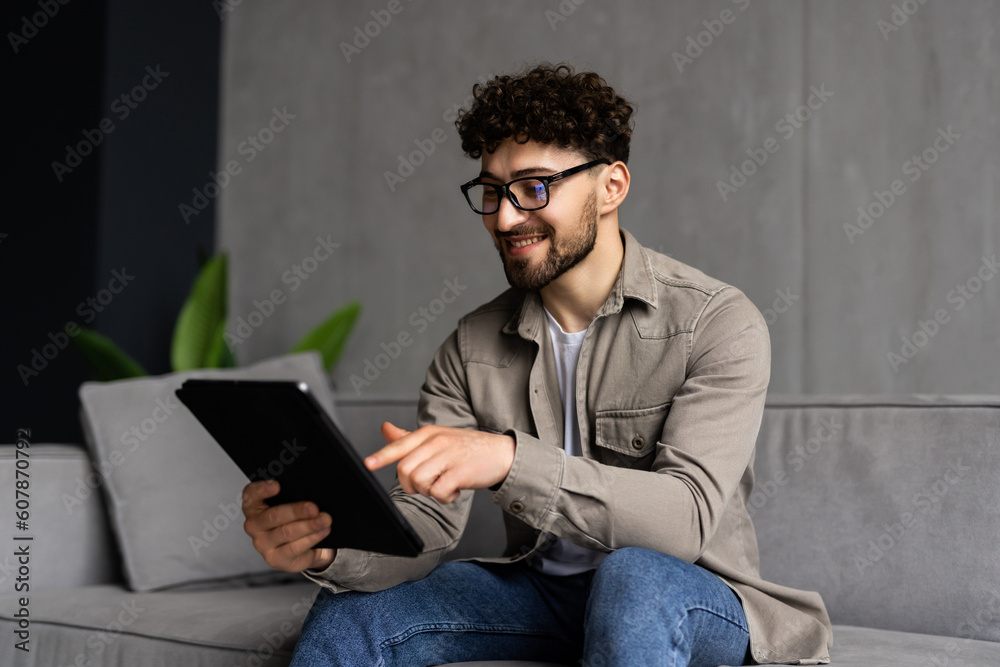 Handsome young man is using a digital tablet and smiling while resting on couch at home