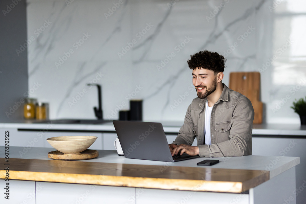 Young man using laptop writing email or message, chatting, working online, sitting in modern kitchen Young man using laptop writing email or message, chatting, working online, sitting in modern kitchen