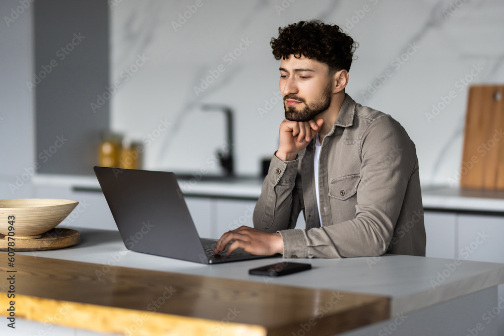 Young man using laptop writing email or message, chatting, working online, sitting in modern kitchen Young man using laptop writing email or message, chatting, working online, sitting in modern kitchen