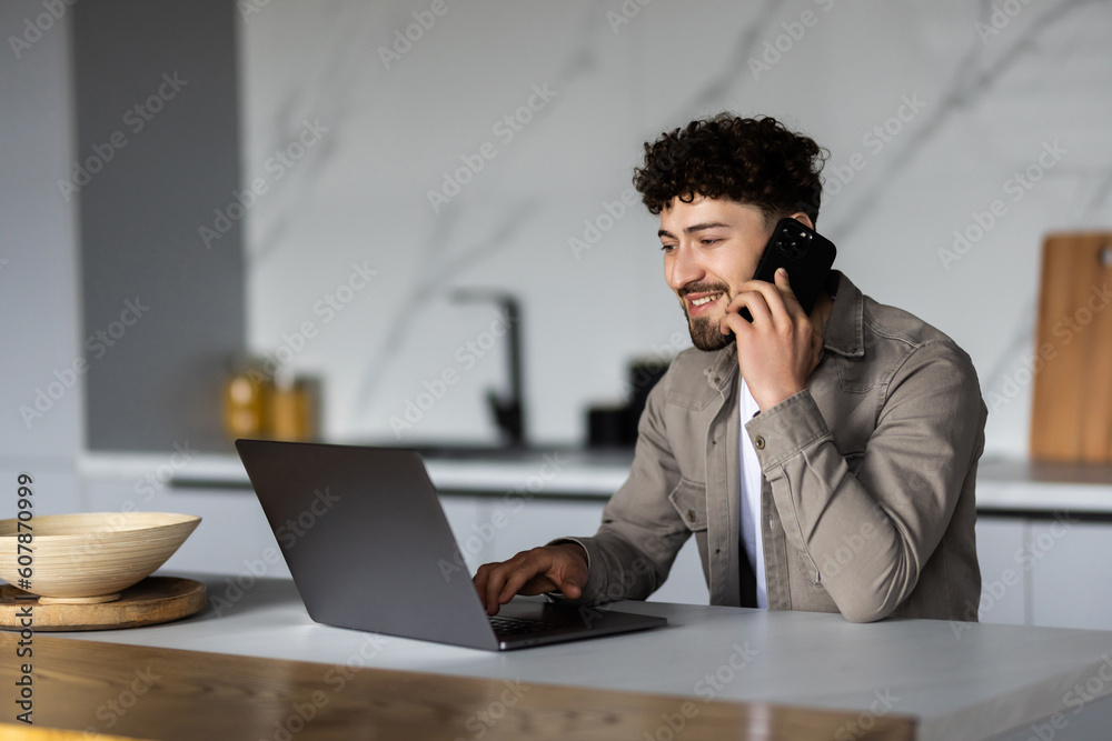 Young man freelancer working remotely at home, looking at laptop and consulting client via cellphone, sitting at kitchen Young man freelancer working remotely at home, looking at laptop and consulting client via cellphone, sitting at kitchen