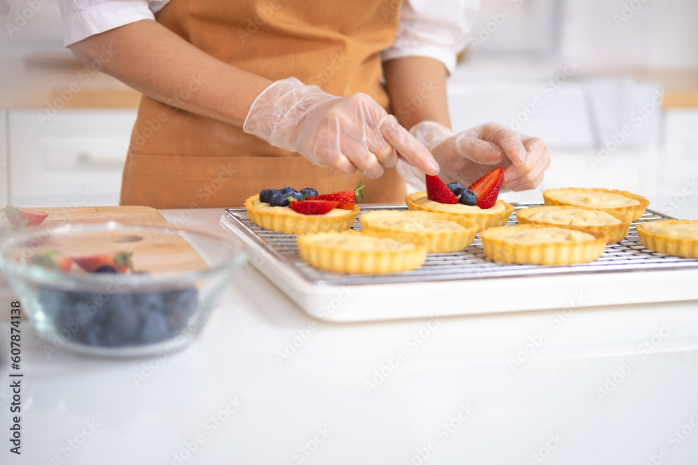 close up of hands in cooking gloves Baker adding blueberries strawberry fresh fruit to a tart on white table in Kitchen. housewife baker wear apron making fruit tart. homemade bakery at home.