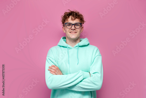 young curly guy student in mint hoodie and glasses stands with his arms crossed on pink isolated background