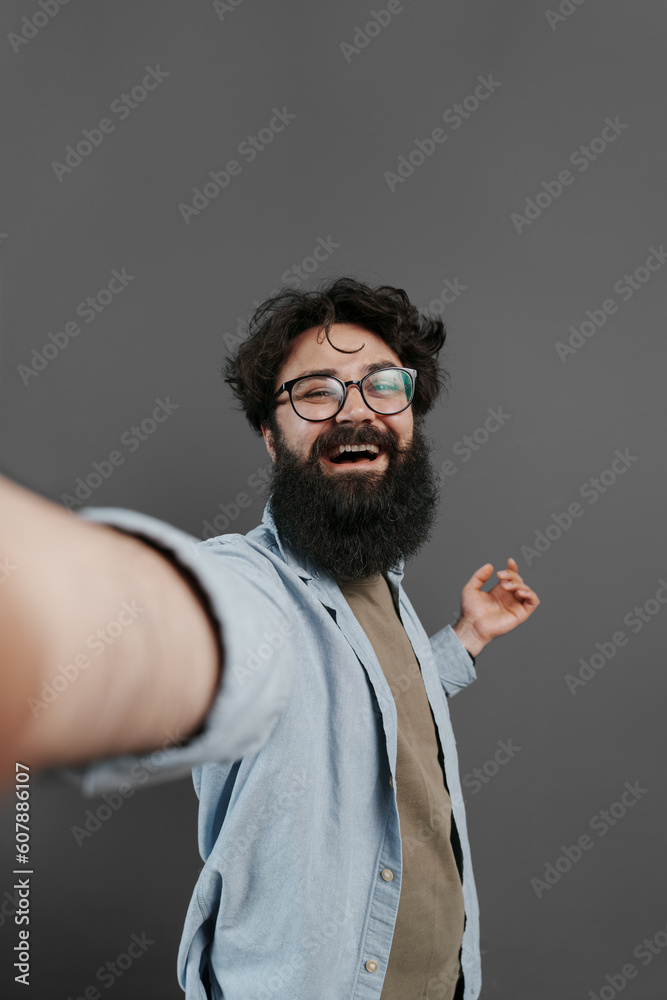Joyful bearded man wearing eyeglasses and denim shirt taking selfie against grey background
