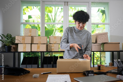 Front view of a young short curly hair Asian female small business owner standing at desk using barcode scanner scanning code on parcel box before shipment in a room with many parcel boxes in the back