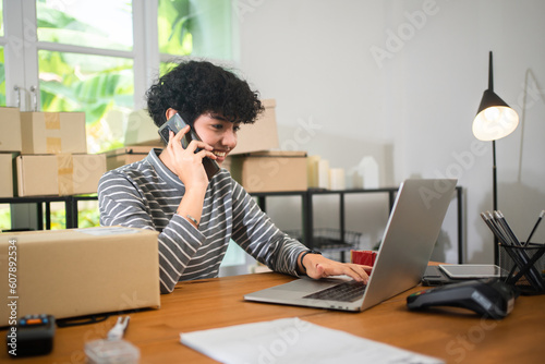 Oblique angle, selective focus of young short curly hair Asian female small business owner sitting at desk, smiling talking on the phone while working on laptop in a room with many parcel boxes behind