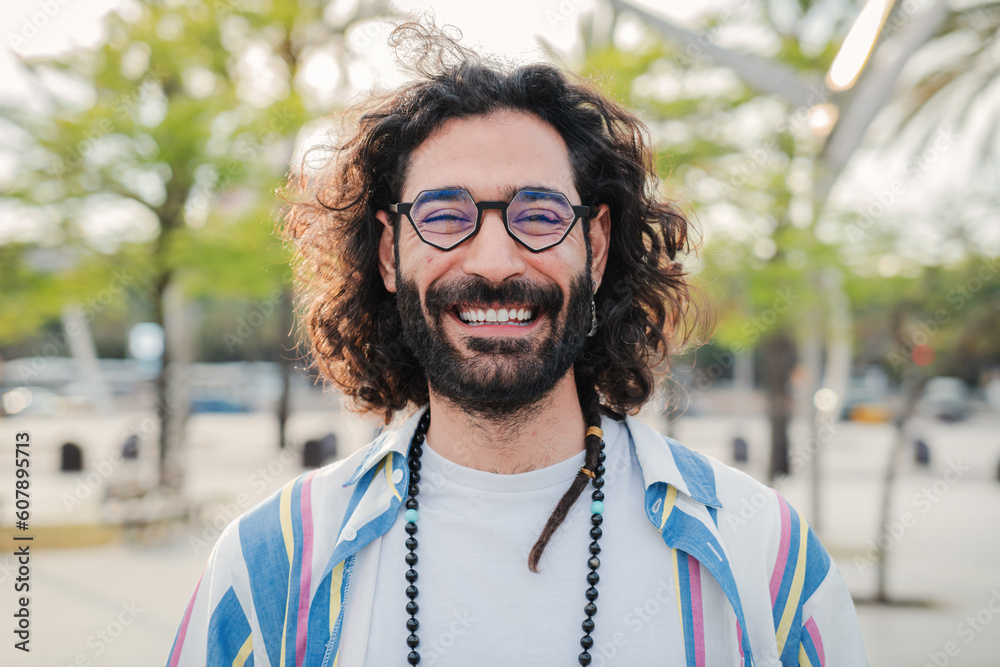Happy young hipster man smiling and looking at camera. Close up individual portrait of hippie guy laughing. Front view of one bearded bohemian male person standing outdoors with cool expression. High Happy young hipster man smiling and looking at camera. Close up individual portrait of hippie guy laughing. Front view of one bearded bohemian male person standing outdoors with cool expression. High
