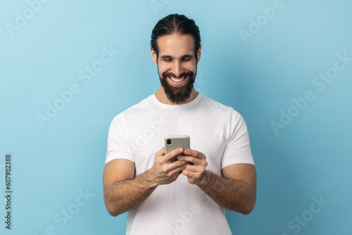 Portrait of handsome positive man bodybuilder with beard wearing white T-shirt using smart phone, looking at device screen, writing post. Indoor studio shot isolated on blue background.