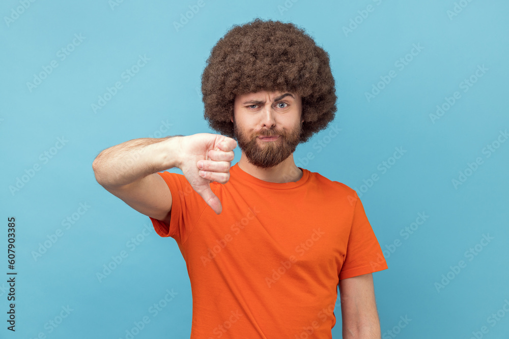 Portrait of displeased man with Afro hairstyle wearing orange T-shirt showing thumbs down dislike gesture, symbol of disagree, giving feedback. Indoor studio shot isolated on blue background.