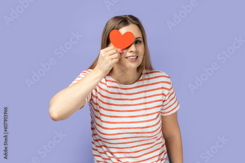 Portrait of positive funny young adult blond woman wearing striped T-shirt holding little red heart and covering her eye behind love symbol. Indoor studio shot isolated on purple background. © khosrork