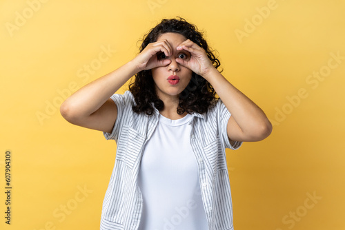 Portrait of woman with dark wavy hair spying, watching unbelievable shocking event, looking through fingers imitating binoculars. Indoor studio shot isolated on yellow background.