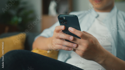 Closeup of Happy attractive Asian man sit on sofa hand holding mobile phone chatting and playing social media. Young male looking at smartphone cellphone browsing internet on sofa at home living room