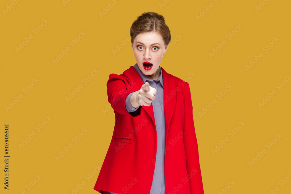 Portrait of shocked astonished woman with red lips standing indicating fore finger to you, being amazed and surprised, choosing you, wearing jacket. Indoor studio shot isolated on yellow background. Portrait of shocked astonished woman with red lips standing indicating fore finger to you, being amazed and surprised, choosing you, wearing jacket. Indoor studio shot isolated on yellow background.
