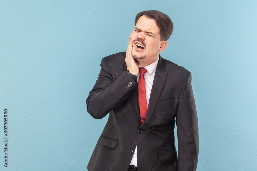 Portrait of man suffers painful toothache, has rotten tooth, needs to visit dentist, keeps eyes closed from pain, wearing black suit with red tie. Indoor studio shot isolated on light blue background. Portrait of man suffers painful toothache, has rotten tooth, needs to visit dentist, keeps eyes closed from pain, wearing black suit with red tie. Indoor studio shot isolated on light blue background.