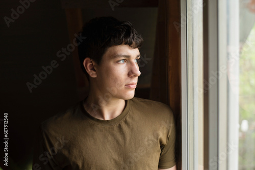 Standing in front of the window and thinking, a young 18-year-old dreams, chooses whether to study or work. Portrait of a charismatic serious guy