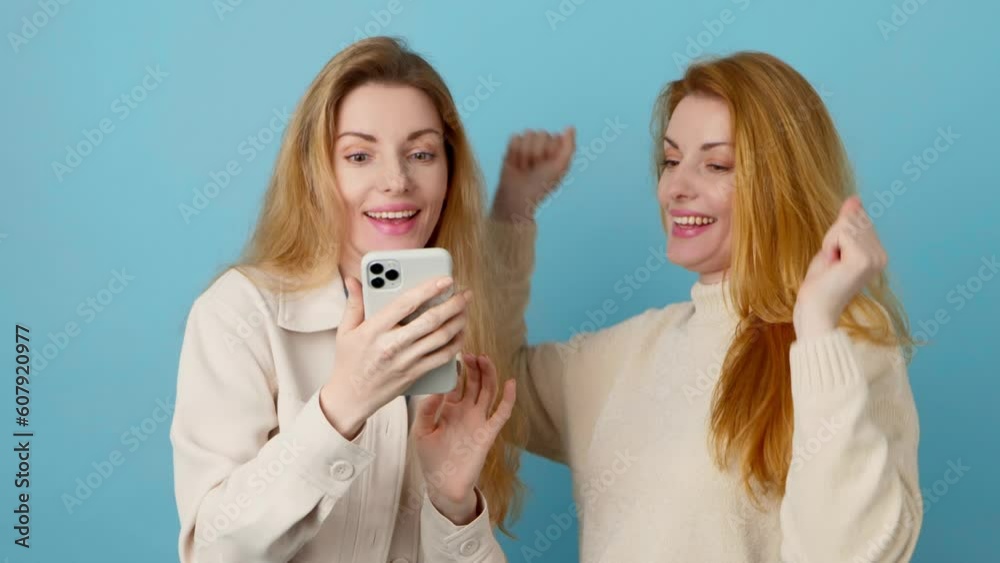 Mobile cell phone, friends say Wow and woman celebration for winning, discount or sale on blue background. Two women sister excited with smartphone for social media, online streaming, news or internet Mobile cell phone, friends say Wow and woman celebration for winning, discount or sale on blue background. Two women sister excited with smartphone for social media, online streaming, news or internet