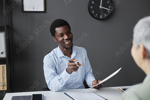 Portrait of young black businessman smiling to colleague at workplace desk in office