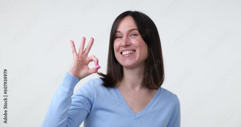 Caucasian woman in blue blouse standing on a white background and flashing a wide smile while making the 'OK' hand gesture. She exudes positivity and approval. Caucasian woman in blue blouse standing on a white background and flashing a wide smile while making the 'OK' hand gesture. She exudes positivity and approval.