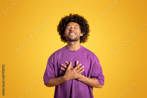Happy satisfied mature black curly man in purple t-shirt with closed eyes presses hands to chest