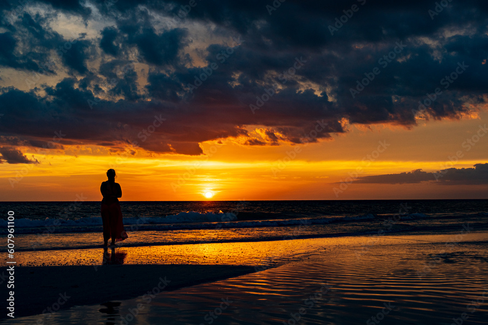 a female stands on the beach while the sunsets below the water on the horizon