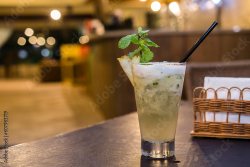 Lemon Caipirinha with sake. Glass of refreshing cold alcoholic beverage. Glass cup decorated with a piece of pineapple and leaves. Bar table with blurred background.