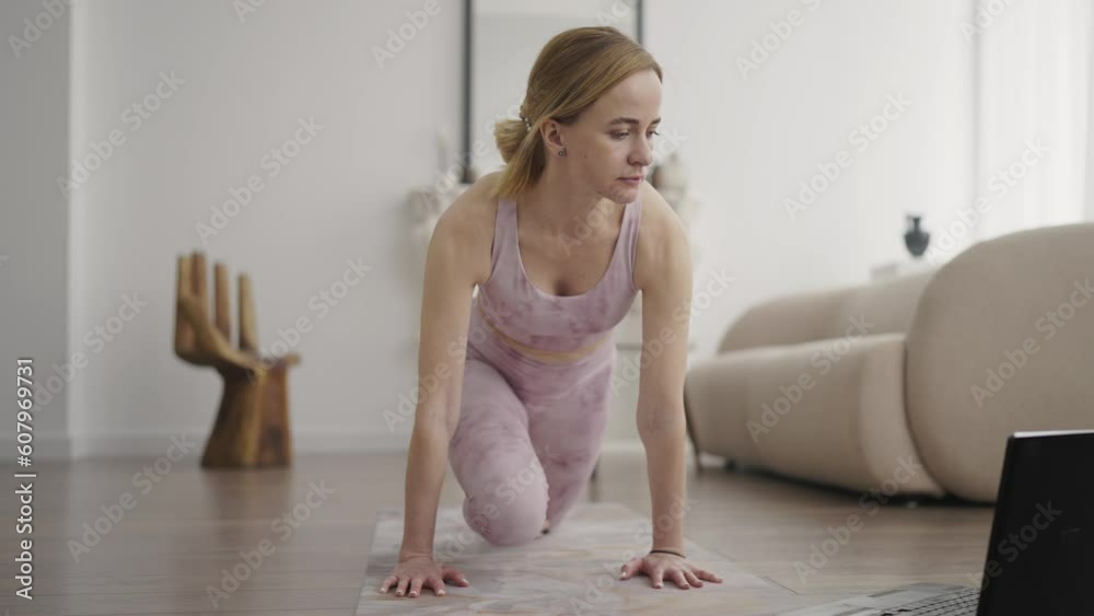 Young girl performs yoga stretching exercise on a rug in a bright room. Girl watches online lesson on laptop. Slow motion Young girl performs yoga stretching exercise on a rug in a bright room. Girl watches online lesson on laptop. Slow motion