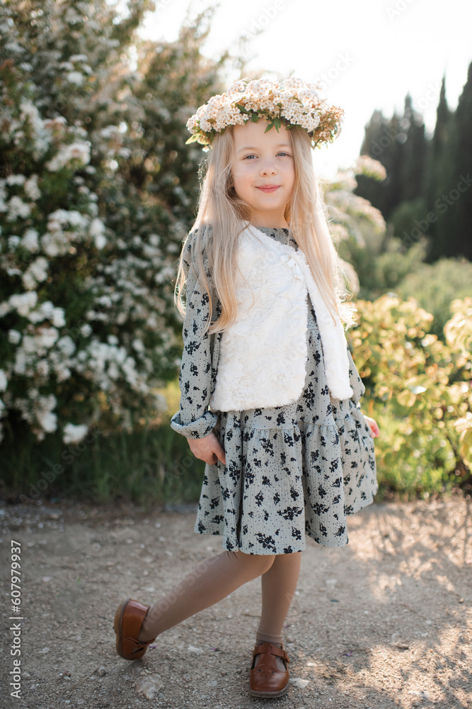 Obraz premium Smiling stylish child girl 4-5 year old wear floral wreath hairstyle and dress standing over flower at background outdoor. Look at camera. Summer season.
