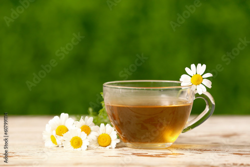 Glass cup of tea with beautiful chamomile flowers on white wooden table outdoors