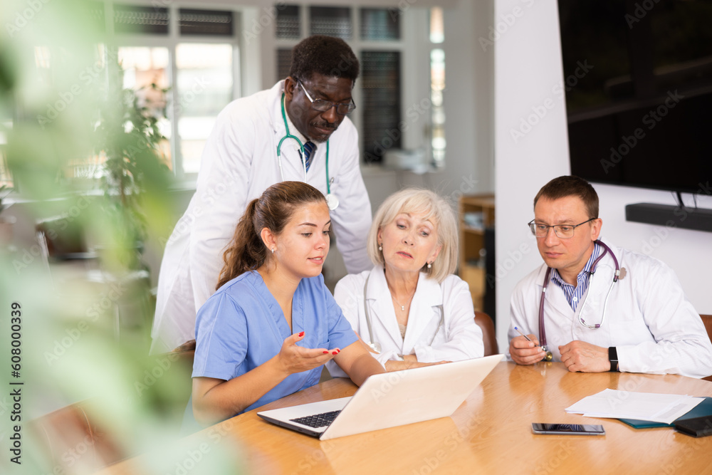 Three physicians sitting and young nurse standing by them looking at the notebook interestedly