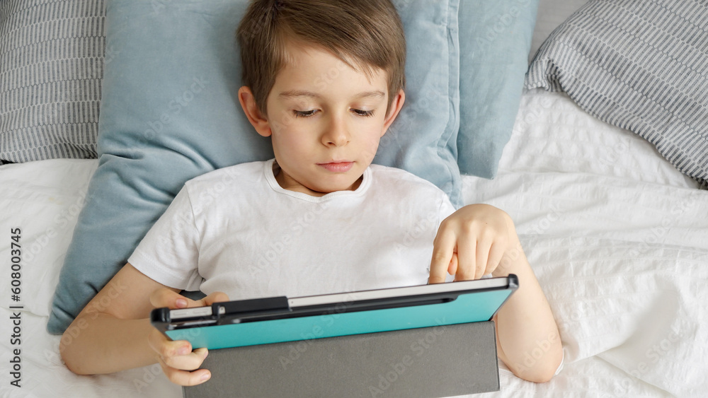 A little boy is lying on his bed and playing with a tablet computer during the day. Child using gadget, education and development. A little boy is lying on his bed and playing with a tablet computer during the day. Child using gadget, education and development.
