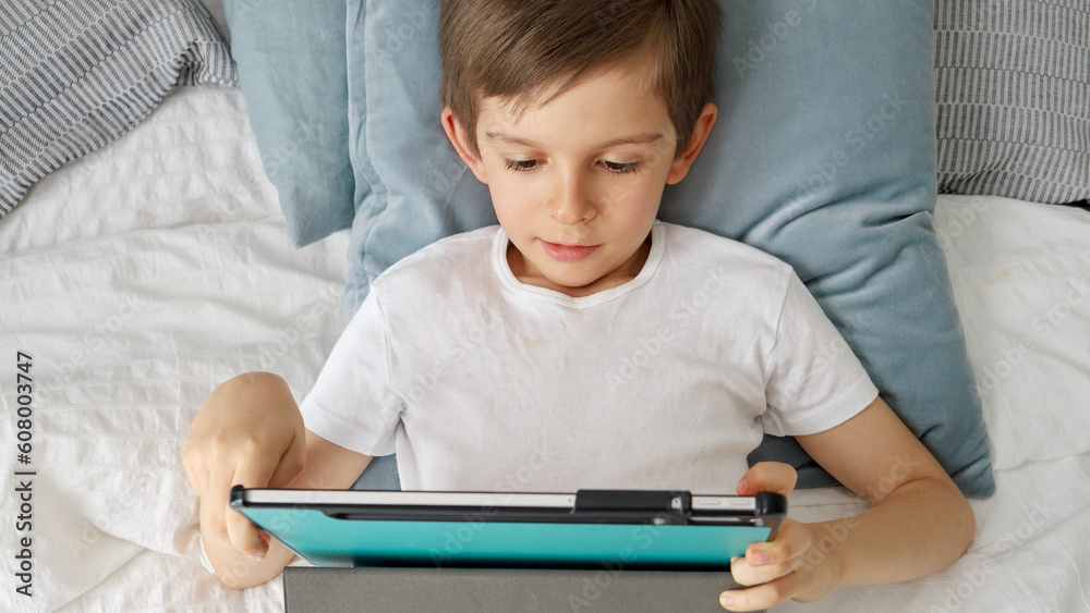 A boy using a tablet computer while lying in bed during the day. Child using gadget, education and development. A boy using a tablet computer while lying in bed during the day. Child using gadget, education and development.