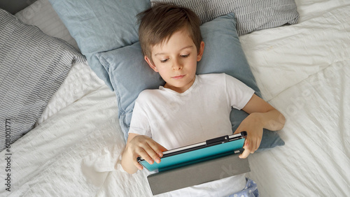 Happy young boy is playing with a tablet computer while lying in bed during the day. Child using gadget, education and development. © Кирилл Рыжов