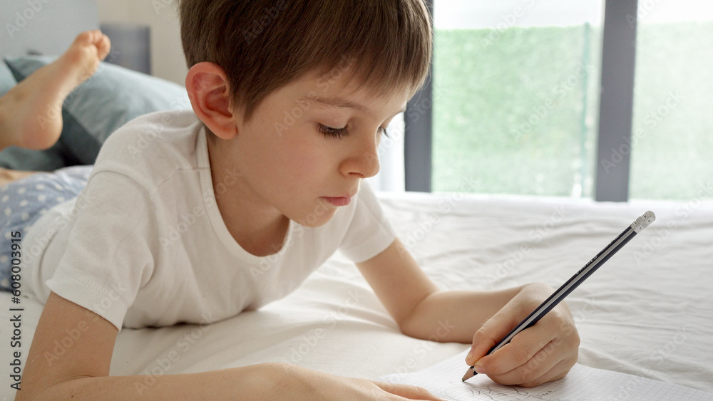 Little boy doing his homework in bed, writing in a notebook with diligence and dedication. Importance of education and child development, remote schooling at home. Little boy doing his homework in bed, writing in a notebook with diligence and dedication. Importance of education and child development, remote schooling at home.